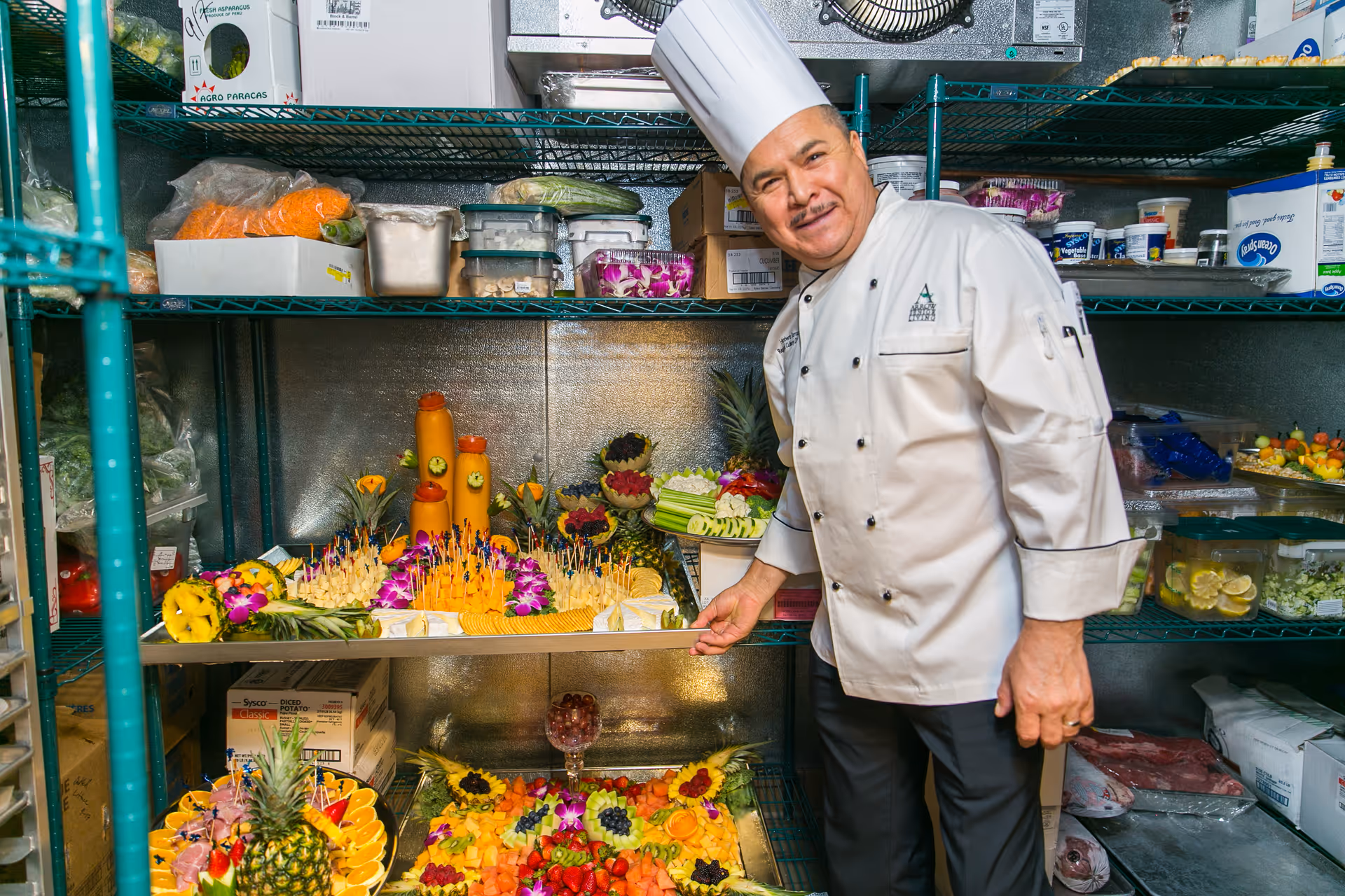 A chef in a white uniform and tall white hat stands in a kitchen storage area, smiling and presenting trays of colorful fruit and vegetable arrangements on metal shelves filled with various food containers and supplies.
