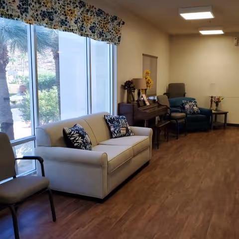 A bright living room area with a large window covered by floral valances. The room features a beige sofa with patterned cushions, a blue armchair, a piano with a lamp and decorative items on top, and a small side table with flowers. The floor is wooden, and the walls are light-colored.