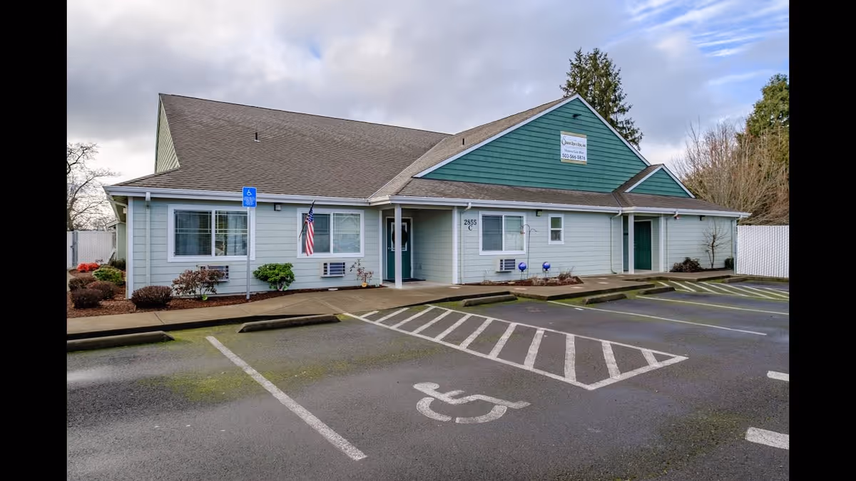 Exterior view of a single-story building with light blue siding and a green gabled roof. The building has several windows with white frames, two green doors, and an American flag mounted near the entrance. There are handicapped parking spaces and a sidewalk in front of the building, with some landscaping including bushes and small trees. The sky is partly cloudy.