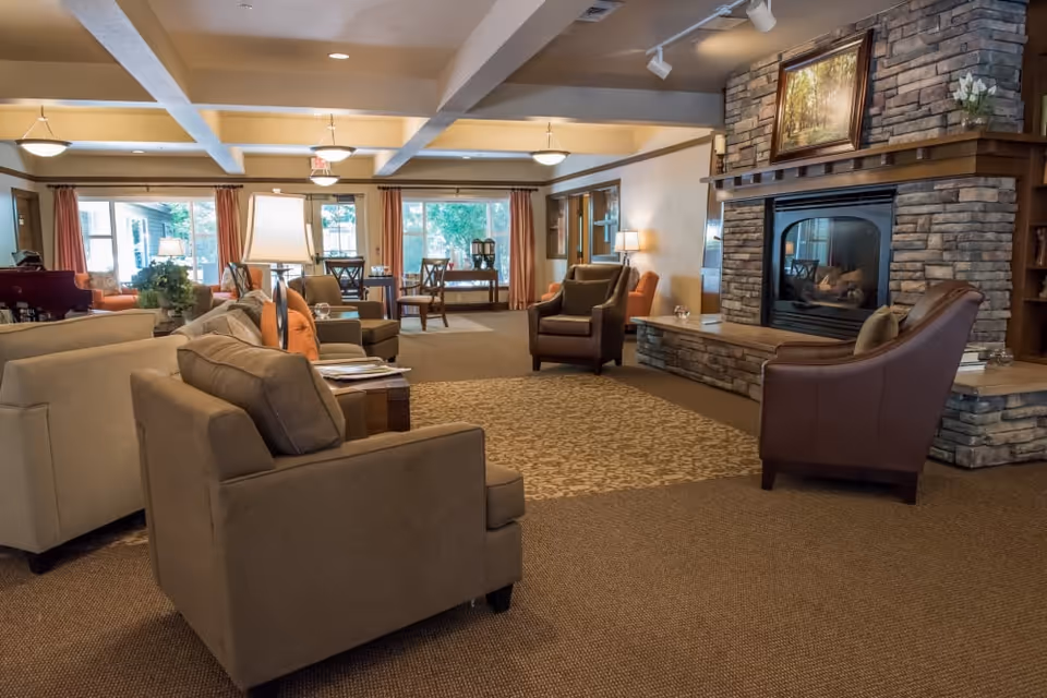 A cozy living room area in a senior living facility with multiple armchairs and sofas arranged around a stone fireplace. The room has warm lighting, large windows with curtains, and a carpeted floor with a patterned rug in front of the fireplace. There are lamps and small tables placed around the seating area, and a framed painting above the fireplace.