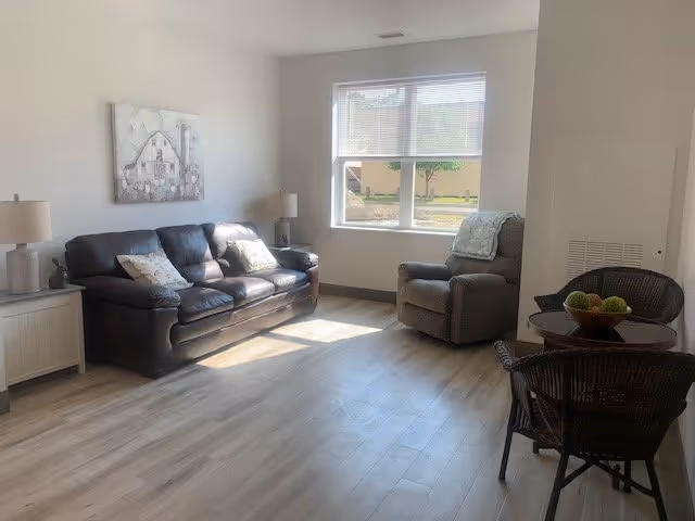 A bright living room with a dark leather sofa, an armchair with a blanket draped over it, a small round table with two wicker chairs, and a window letting in natural light. The room has light-colored wooden flooring and neutral-colored walls with a painting above the sofa.