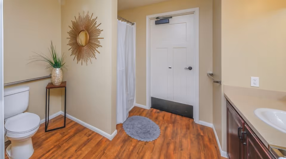 A clean bathroom with a white toilet, a small wooden table holding a decorative vase with green plants, a sunburst mirror on the beige wall, a white shower curtain, a gray circular rug on the wooden floor, and a white door with a metal kick plate. There is also a sink with a beige countertop and dark wood cabinets.