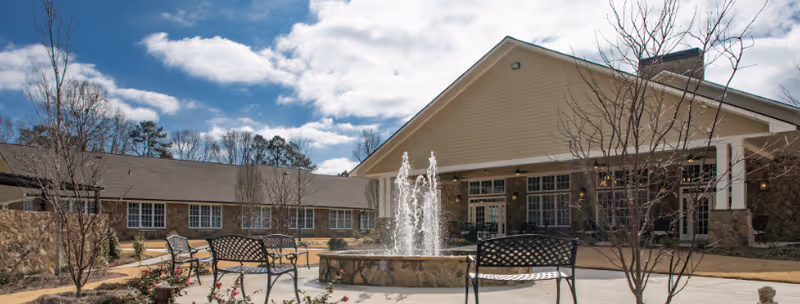 Outdoor courtyard area at Manor Lake Assisted Living and Memory Care - BridgeMill featuring a central stone fountain with water jets, surrounded by benches and young trees under a partly cloudy sky.