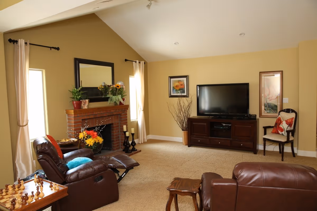 A cozy living room with beige walls and carpet, featuring two brown leather recliners, a wooden side table, a brick fireplace decorated with plants and flowers, a large flat-screen TV on a wooden TV stand, framed artwork on the walls, and two windows with cream-colored curtains.