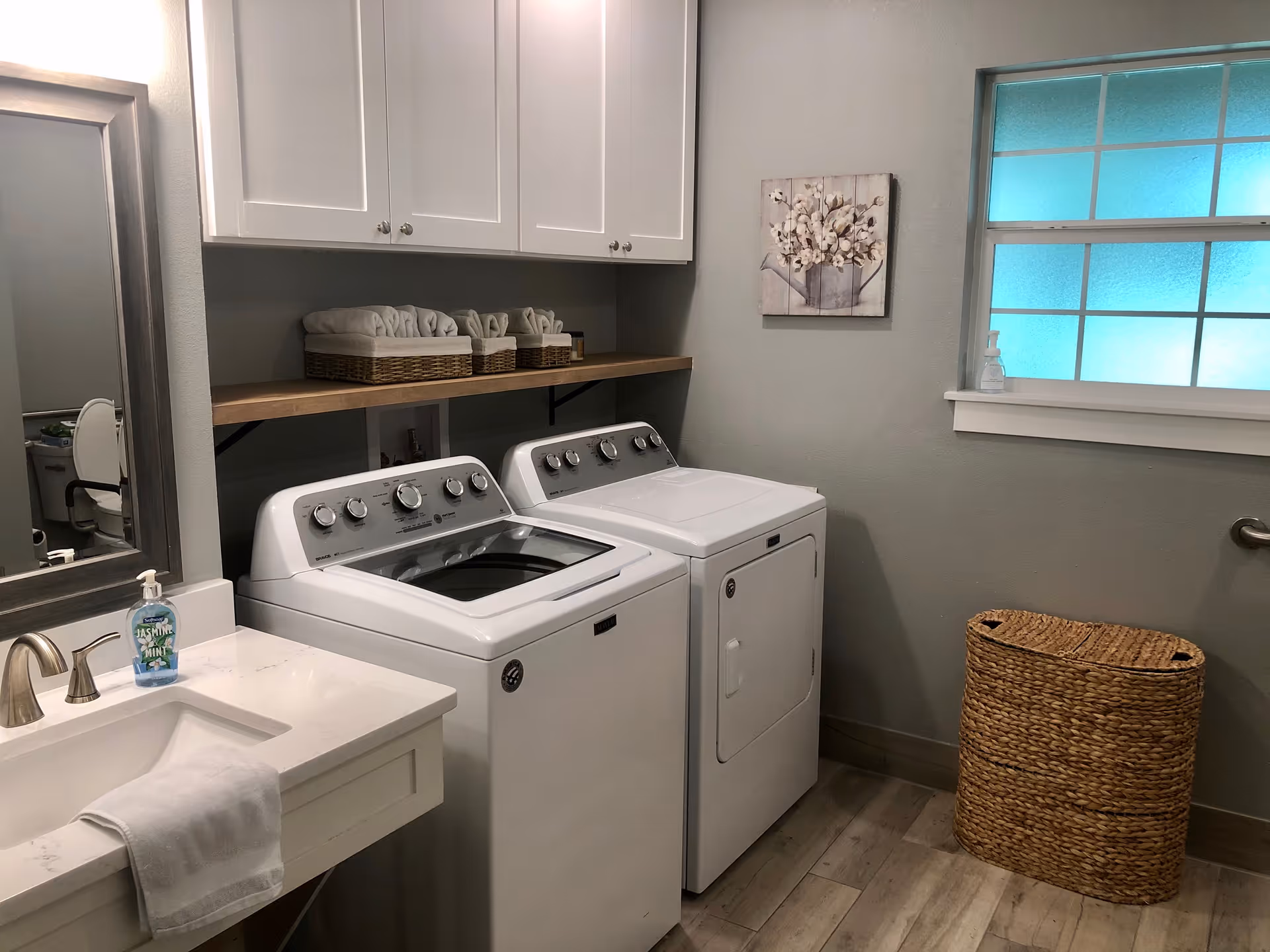 A laundry room with a white washing machine and dryer side by side. Above them is a wooden shelf holding baskets with folded towels. To the left is a white sink with a faucet and a bottle of hand soap. A large mirror is mounted on the wall above the sink. On the right side, there is a window with frosted glass and a woven laundry basket below it. The walls are painted light gray and the floor has wood-like tiles.