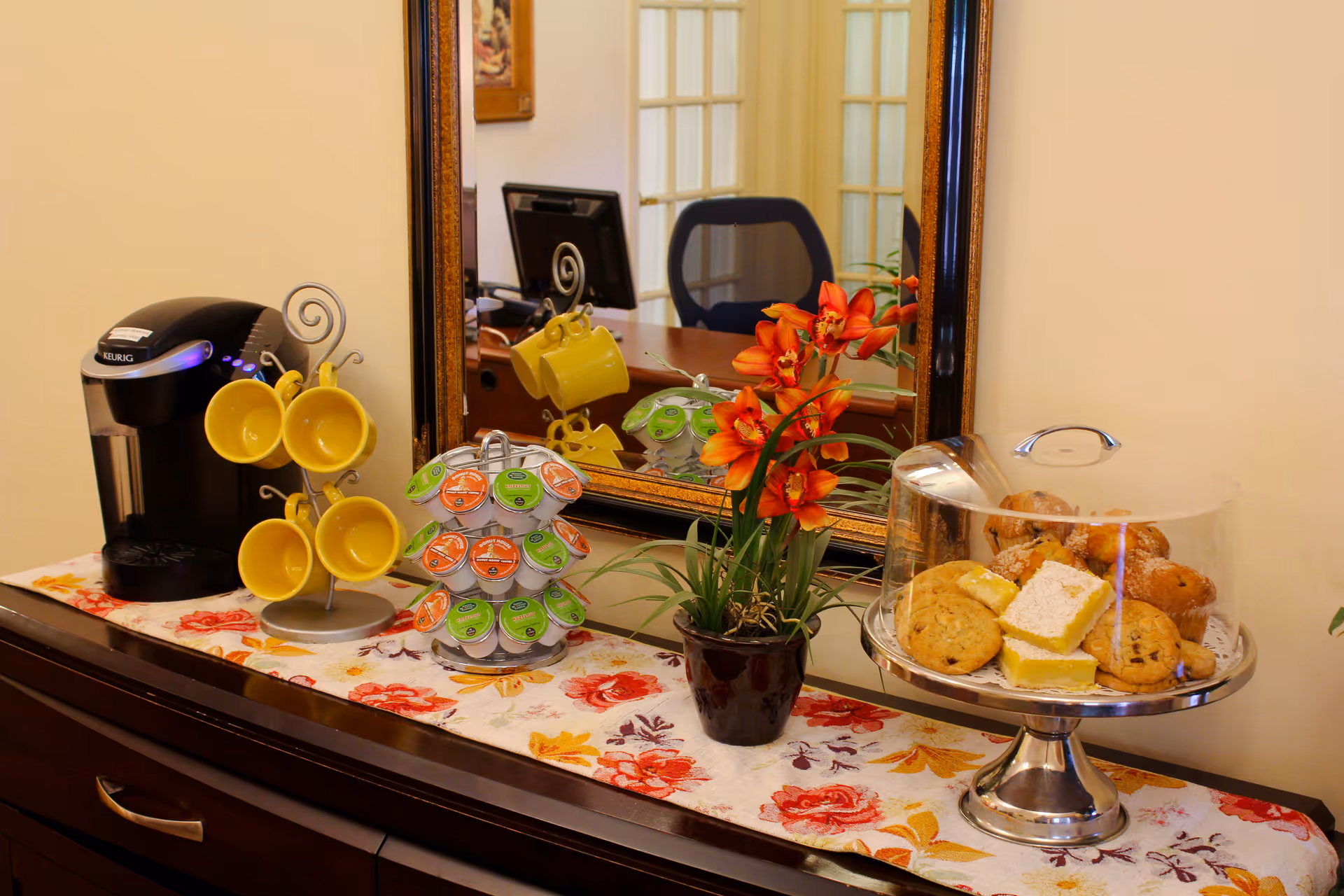 A coffee station setup on a wooden sideboard with a Keurig coffee maker, yellow coffee mugs hanging on a metal stand, a rotating holder with coffee pods, a small potted plant with orange flowers, and a glass dome covering assorted cookies and pastries. A large mirror reflects part of an office chair and desk in the background.