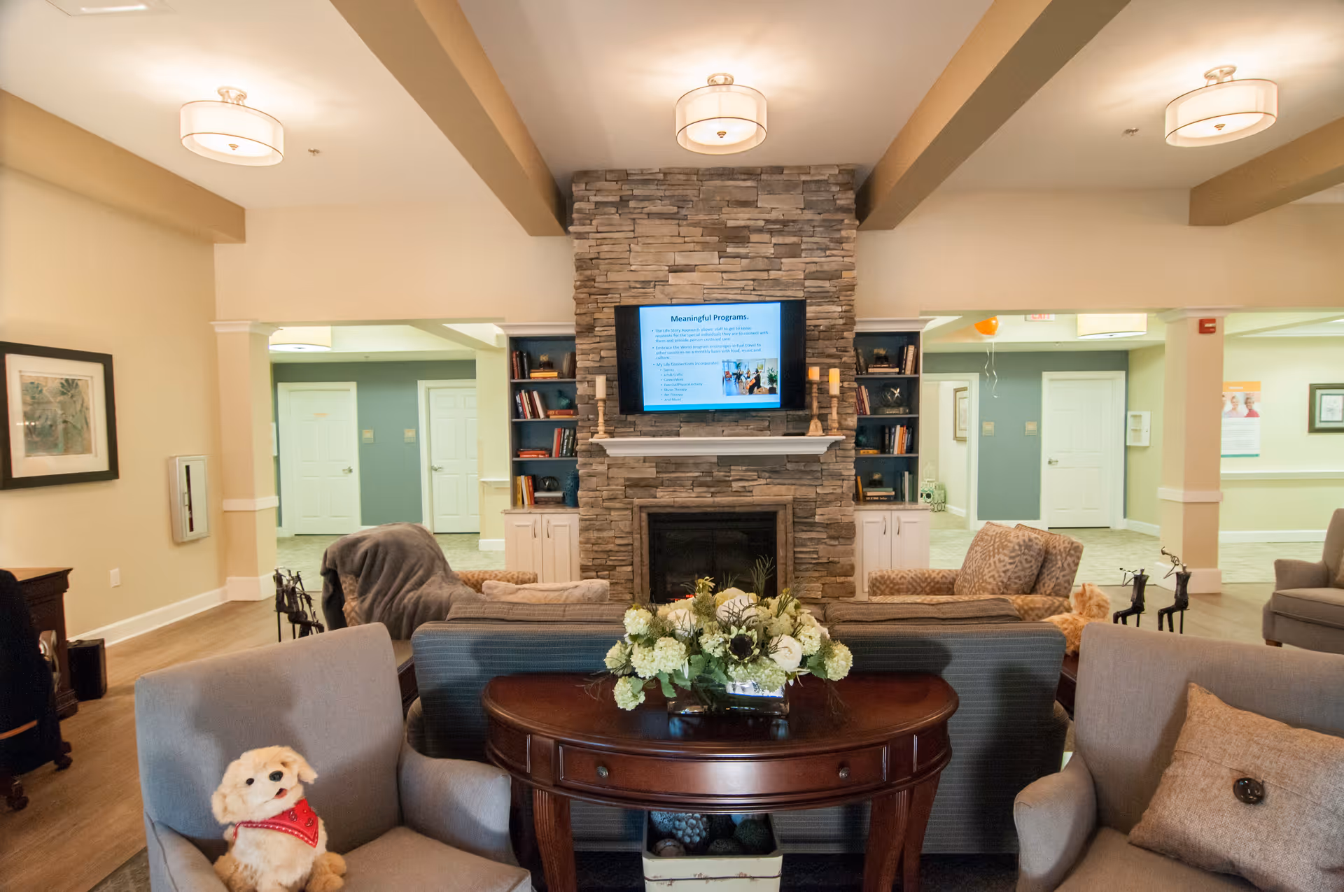Comfortable senior living lounge with sofas and chairs arranged around a stone fireplace and wall-mounted TV.