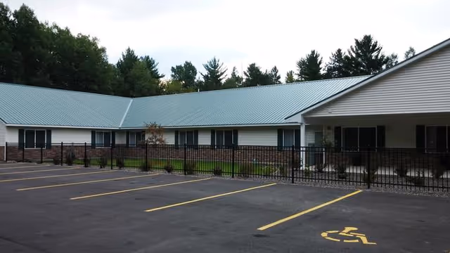 Exterior view of a single-story building with a green metal roof, white siding, and brick lower walls. The building is surrounded by a black metal fence and has a parking lot in front with marked parking spaces, including a handicapped parking spot. Trees are visible in the background under a cloudy sky.