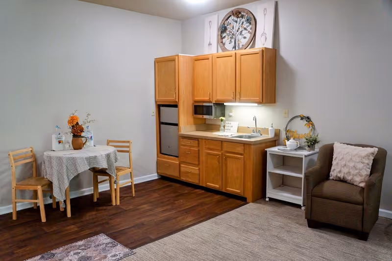A small studio living area with a kitchenette featuring wooden cabinets, a sink and microwave, a round table with two chairs, and an armchair with a pillow.
