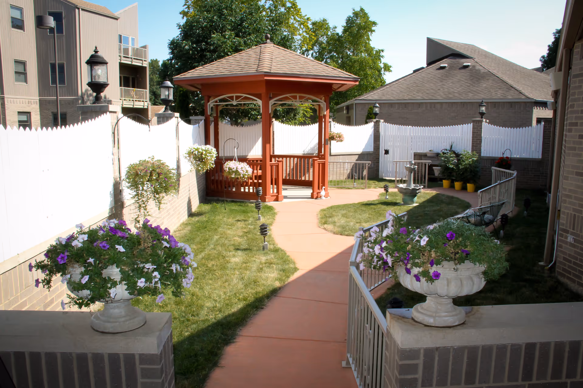 Courtyard with a central red wooden gazebo, paved walkway, grassy areas, and flower-filled planters between brick buildings.