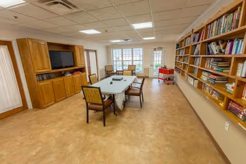 Well-lit communal activity room with a central table and chairs, bookshelves lining one wall, a TV cabinet, and large windows.