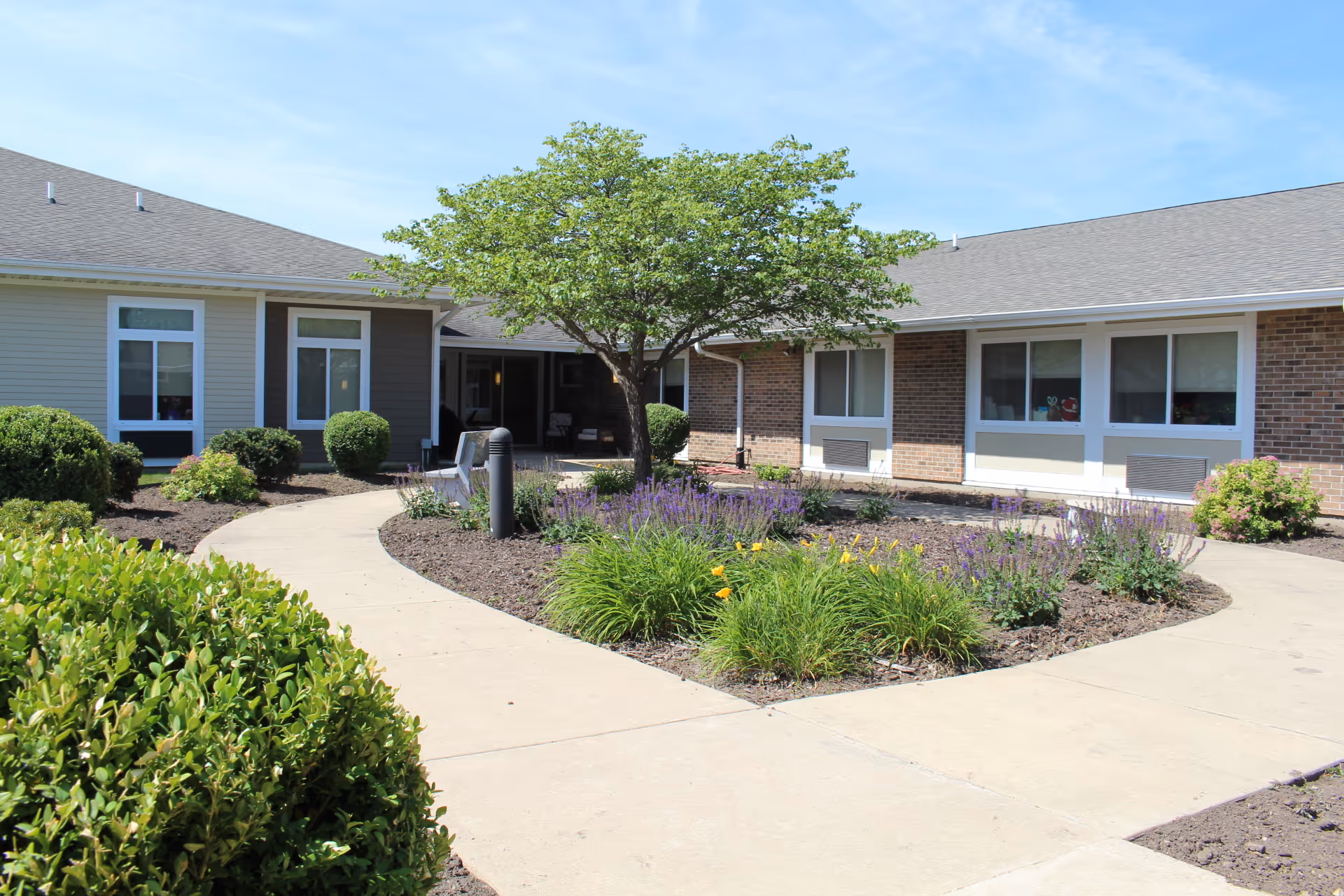 Outdoor courtyard area of a senior living facility with a paved walkway surrounding a garden bed with green shrubs, purple and yellow flowers, and a tree in the center. The building has a mix of brick and siding exterior with several windows and a covered seating area in the background.