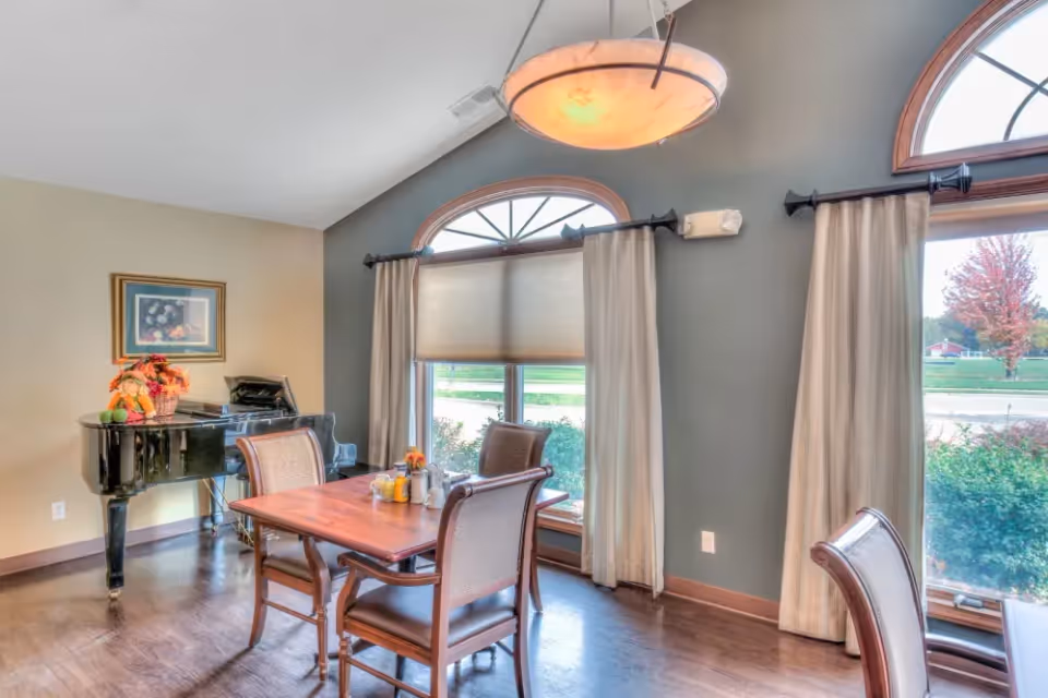 A bright dining area with a wooden table and four cushioned chairs. Large windows with beige curtains and arched transoms let in natural light. A black grand piano with a decorative basket and framed artwork is positioned against the wall. A modern hanging light fixture is above the table.