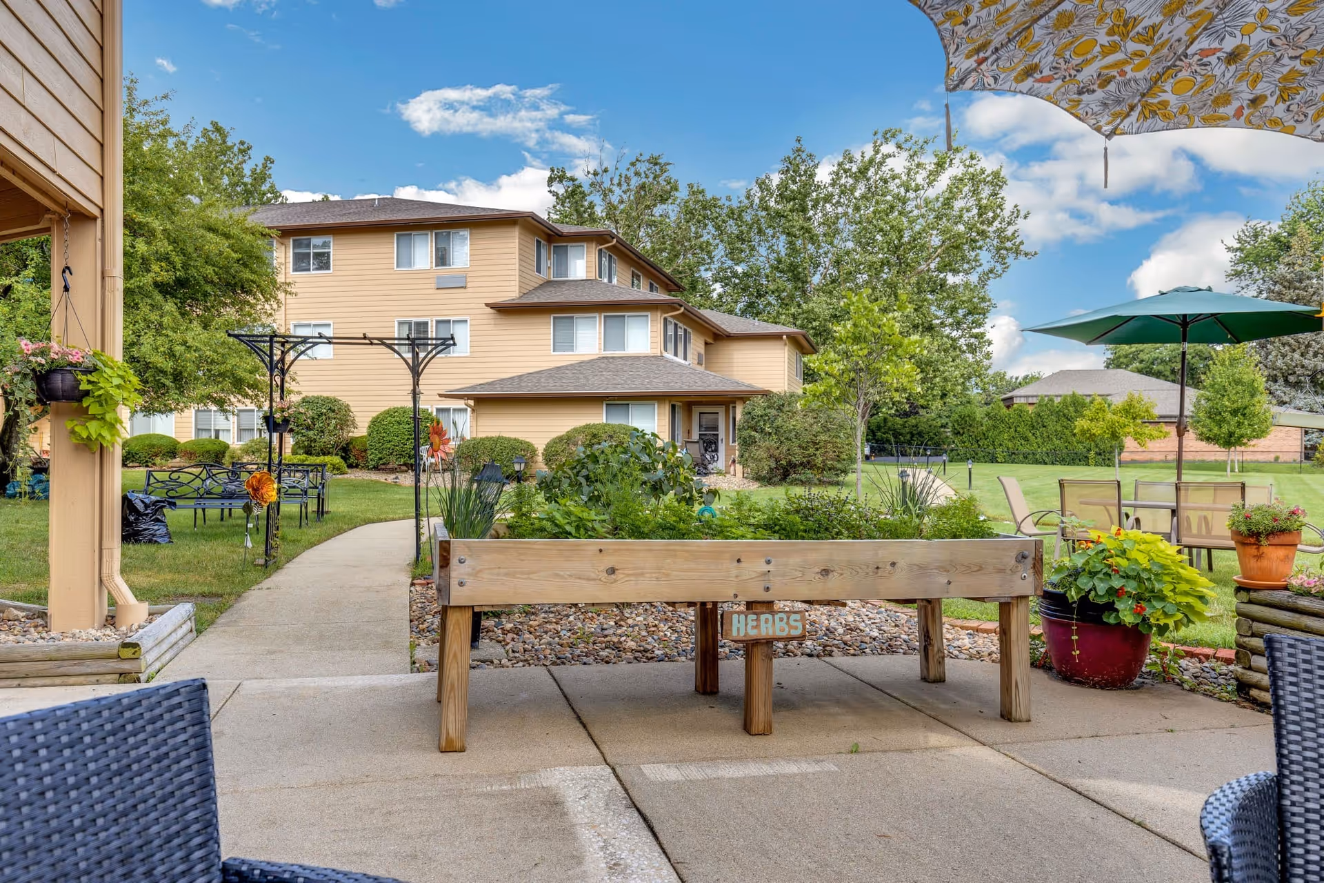 Outdoor garden area at Holiday Blair House featuring a raised wooden herb planter labeled 'HERBS', patio seating with umbrellas, a paved walkway, green lawn, trees, and a multi-story beige building in the background under a partly cloudy blue sky.