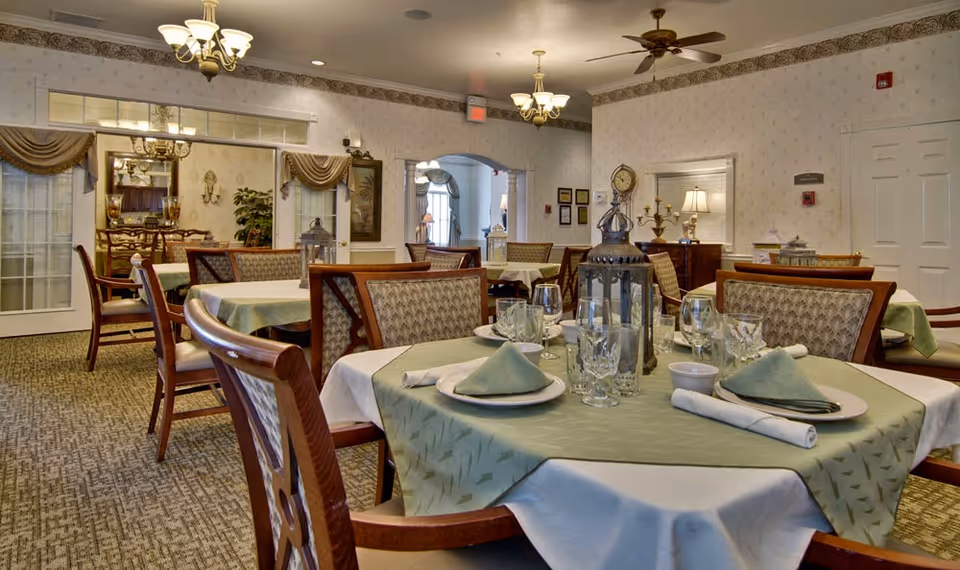 Elegant dining room with multiple set tables, chairs, and decorative centerpieces in a senior living facility.