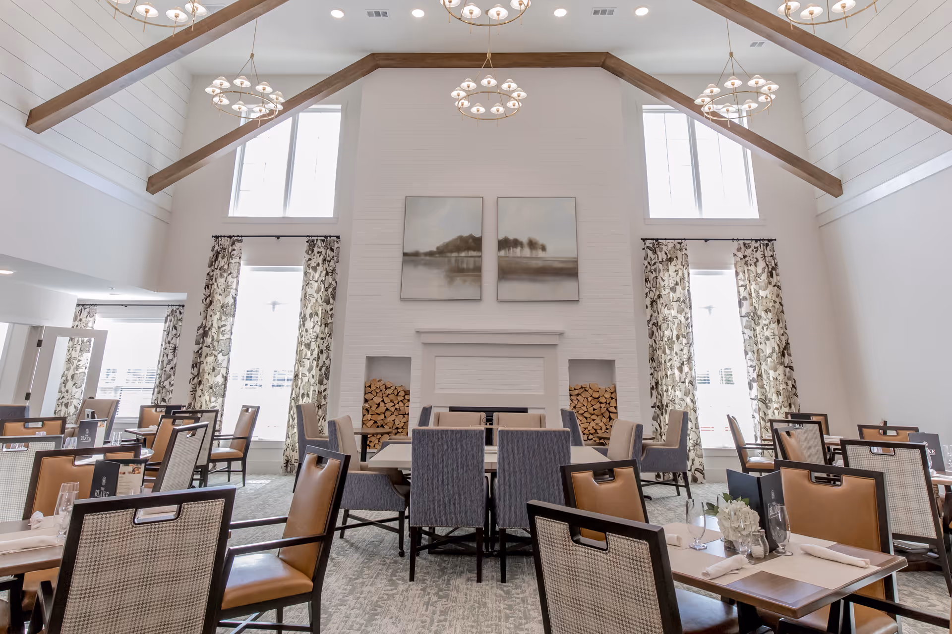A bright and spacious dining room with high vaulted ceilings featuring wooden beams and modern chandeliers. The room has large windows with patterned curtains allowing natural light to fill the space. There are multiple tables and chairs arranged neatly, with place settings including napkins and glassware. A white brick fireplace with stacked firewood on both sides and two landscape paintings above it is the focal point of the room.