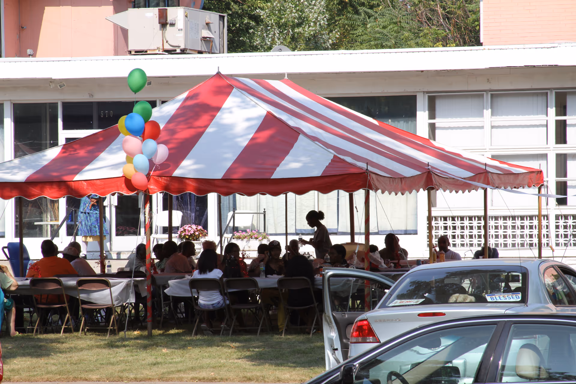Red-and-white striped party tent with people seated at tables beneath it, balloons on a pole and parked cars in front of a building.