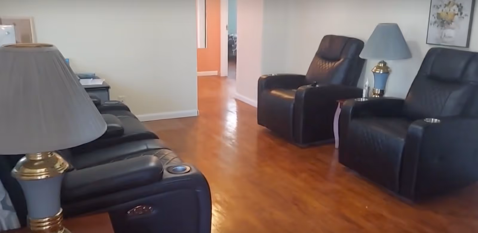 A living room with three black leather recliner chairs, two table lamps with blue shades and gold bases, a small side table, and a framed floral picture on the wall. The floor is wooden, and there is a hallway visible in the background.