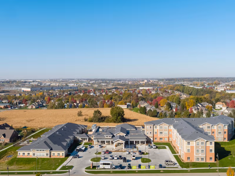 Aerial view of Cedarhurst Senior Living of La Vista facility showing multiple connected buildings with parking lots, surrounded by residential houses and open fields under a clear blue sky.