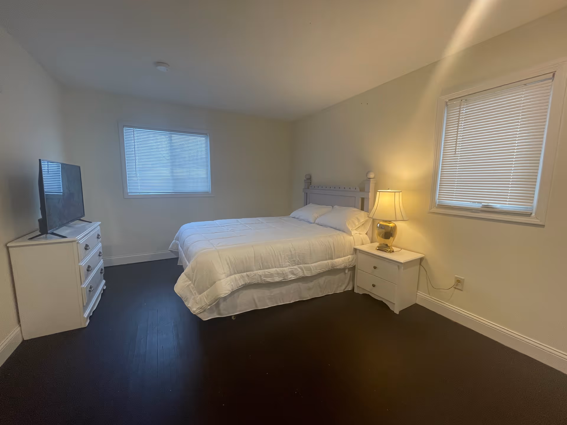 Minimal bedroom with a bed, white nightstands (one with a lamp), a dresser with a TV, two windows, and dark hardwood flooring.