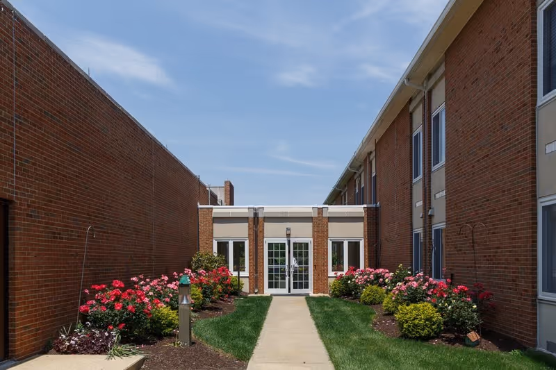 A walkway leading to a glass door entrance between two brick buildings. The path is bordered by well-maintained flower beds with blooming pink and red flowers and green shrubs under a blue sky.