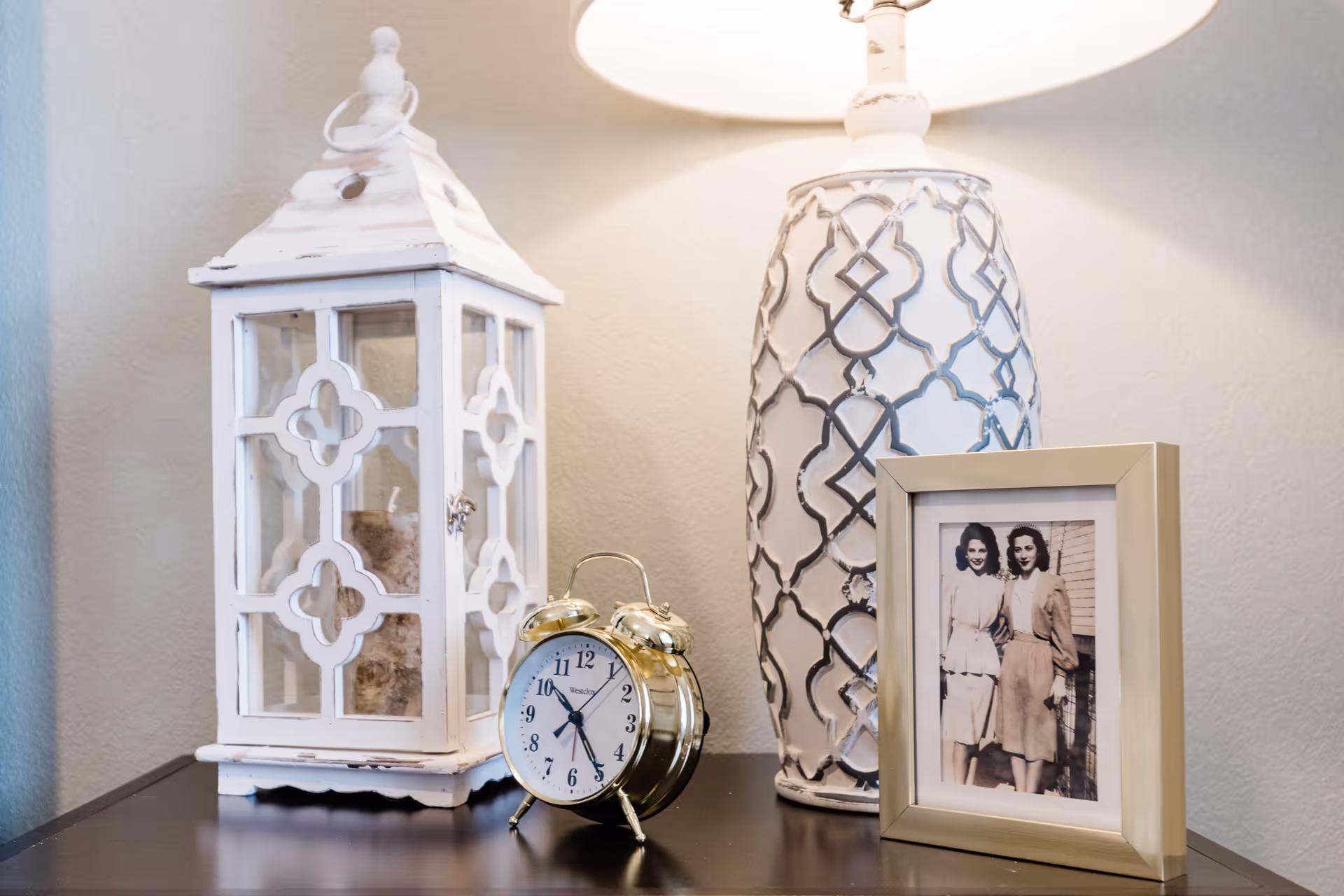 A decorative arrangement on a dark wooden surface featuring a white distressed lantern with a candle inside, a vintage-style gold alarm clock showing the time as 6:27, a white lamp with a patterned base, and a framed black-and-white photograph of two women standing side by side.