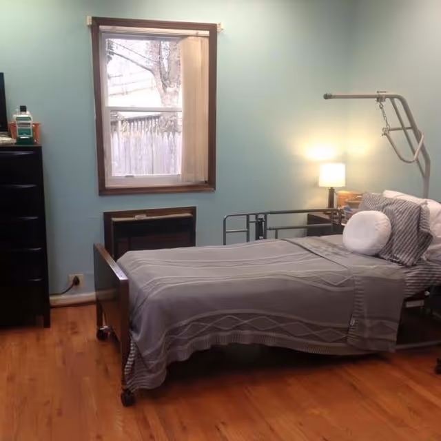 A bedroom with a hospital-style bed covered with a gray blanket and pillows. There is a bedside table with a lamp and some items on it, a window with a view of a wooden fence and tree outside, a black dresser with a bottle on top, and light blue walls with wooden flooring.