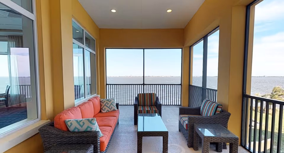 A screened-in porch area with a view of a large body of water. The porch has yellow walls and contains outdoor wicker furniture including a coral-colored sofa with patterned pillows, two striped armchairs, and two glass-top tables. Large windows and screened sections allow natural light and a clear view of the water and sky.