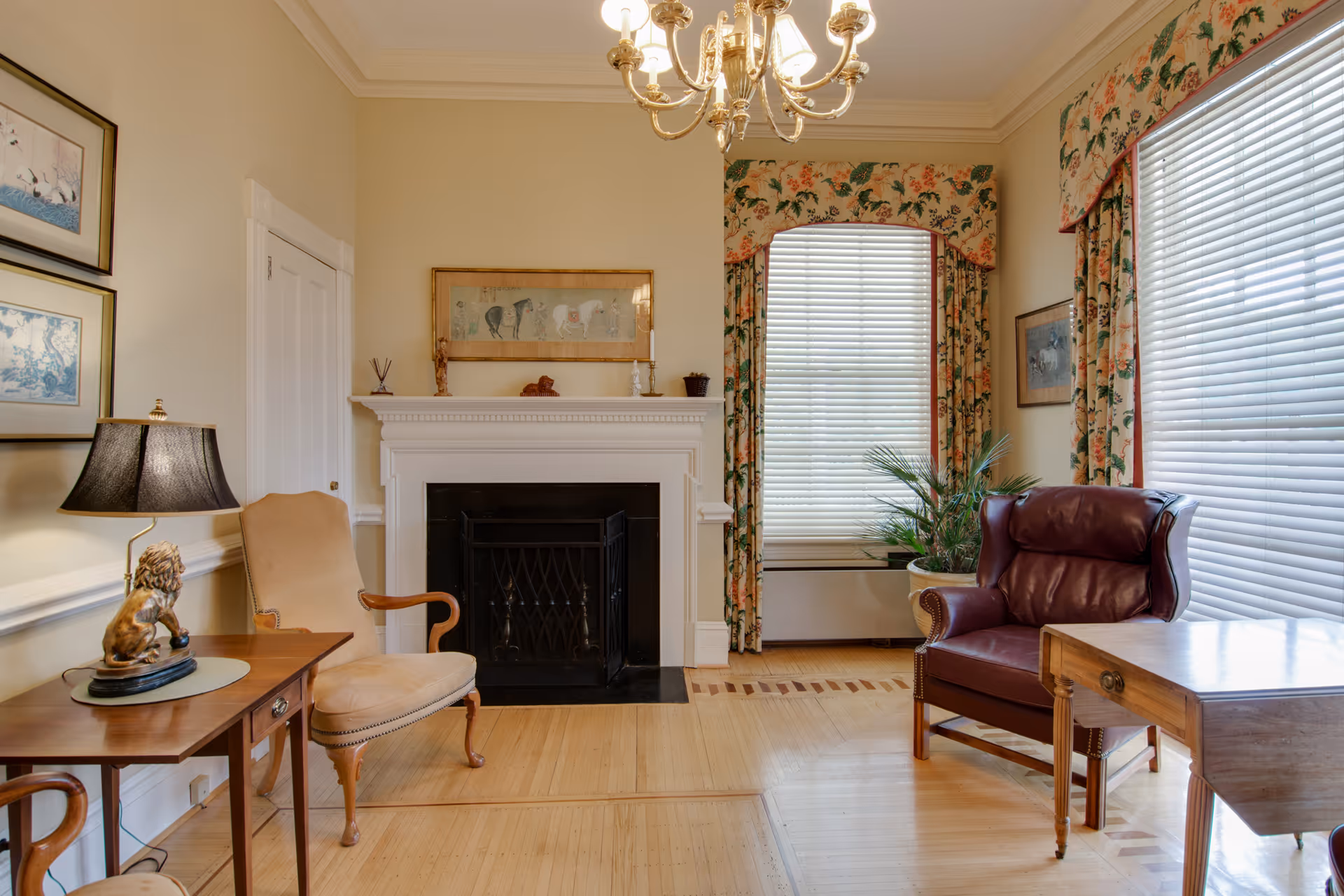 A cozy living room with a white fireplace, two large windows with floral curtains and blinds, a beige armchair, a dark red leather armchair, a wooden side table with a lamp featuring a lion figurine, a wooden table, and framed artwork on the walls.