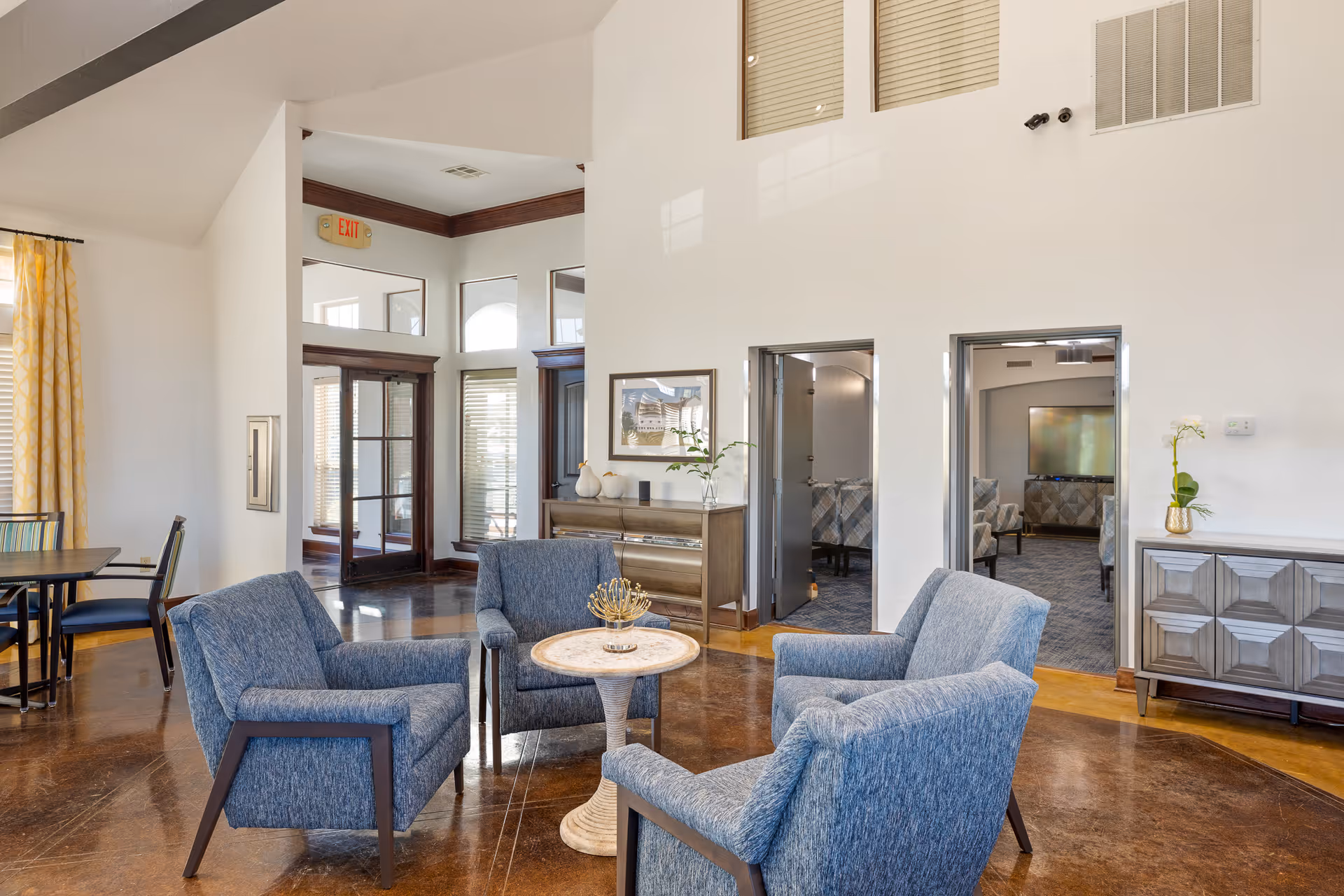 A bright and spacious living room area in a senior living facility with four blue upholstered armchairs arranged around a small round table. The room has polished brown floors, white walls, and large windows with yellow patterned curtains. There are two open doorways leading to another room with additional seating and a large TV. A wooden sideboard with decorative items and a framed picture is against the wall.