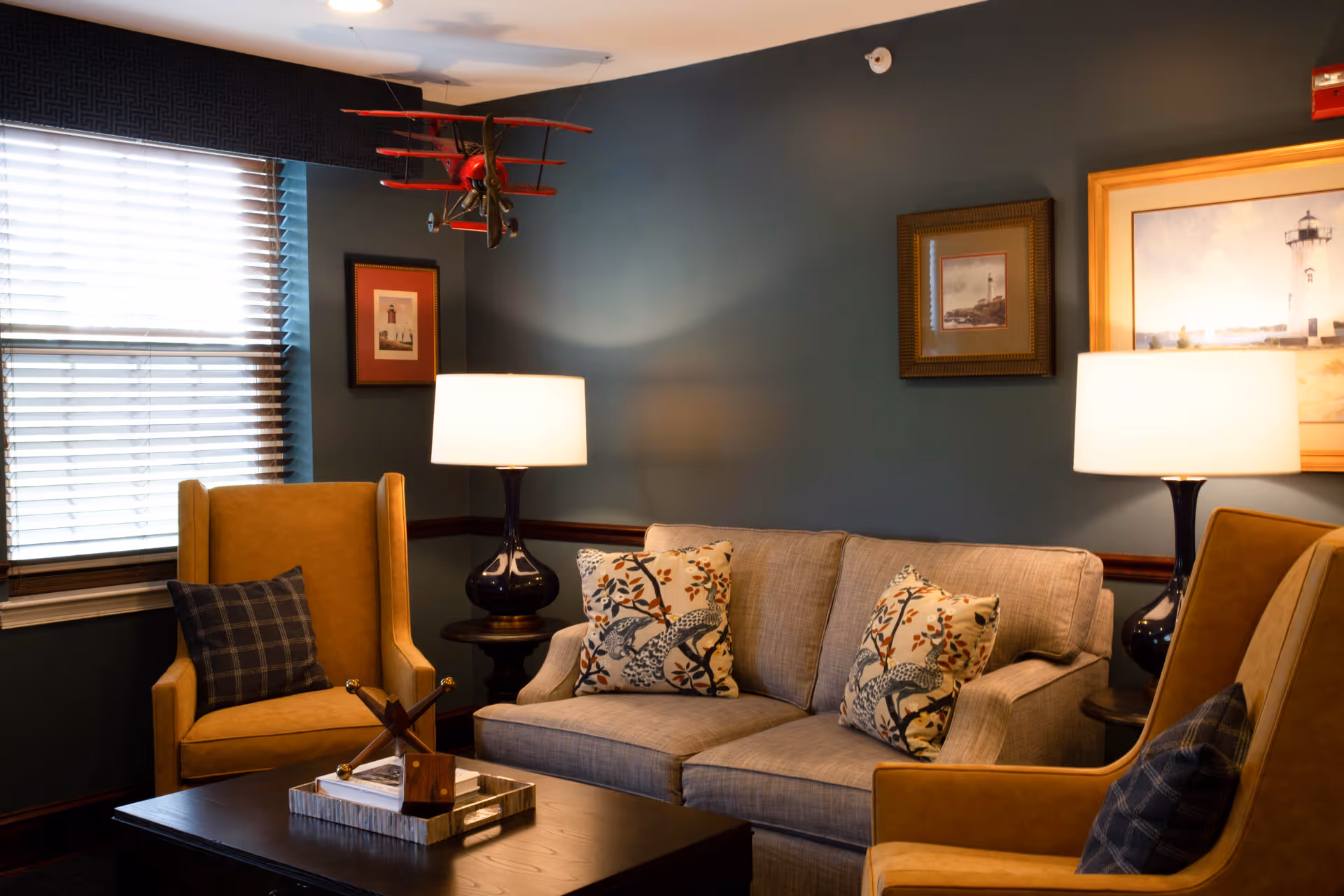 A cozy living room with a beige sofa adorned with patterned pillows, two mustard yellow armchairs with dark plaid cushions, two table lamps with white shades on either side of the sofa, a dark wooden coffee table with decorative items, framed artwork on the walls, and a red model airplane hanging from the ceiling near a window with blinds.