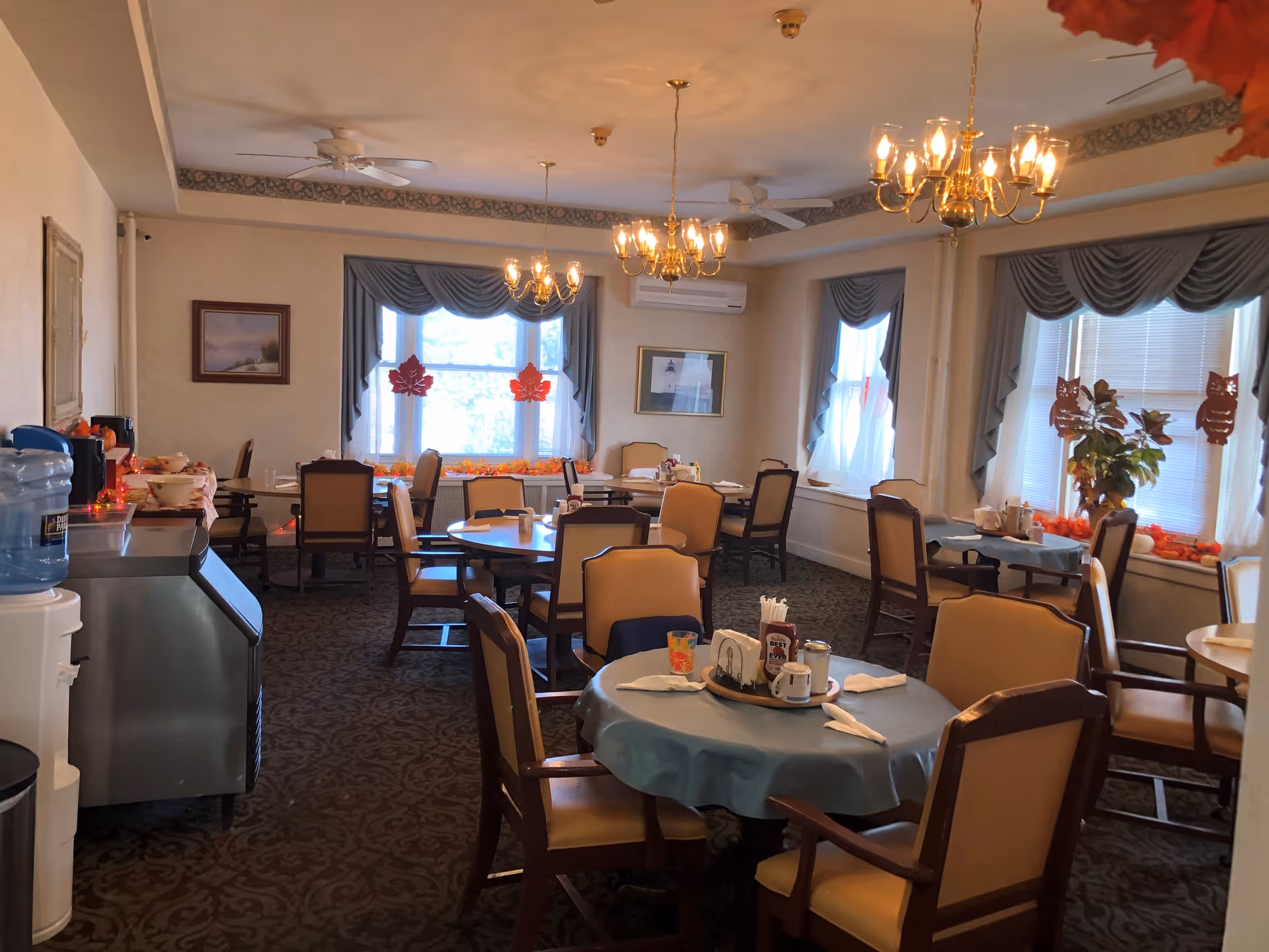 A dining room with several round and rectangular tables covered with tablecloths and set with napkins, cups, and condiments. The room has multiple windows with blue curtains and autumn-themed decorations such as leaves and owls. There are chandeliers hanging from the ceiling, and a water cooler and ice machine are visible on the left side.