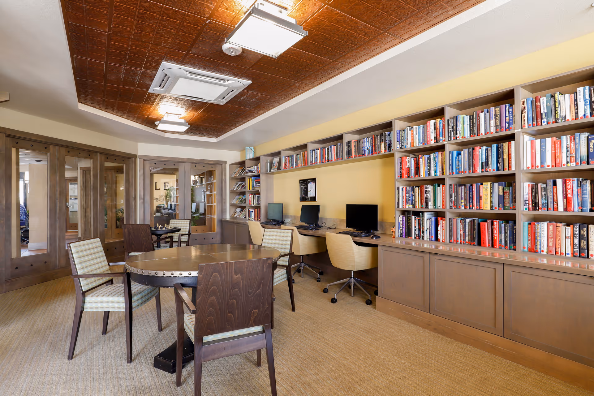 A cozy library and computer area in a senior living facility featuring wooden bookshelves filled with books, a long desk with three computer workstations and chairs, and a round wooden table with four chairs in the center of the room. The ceiling has a decorative wooden panel design with modern light fixtures.