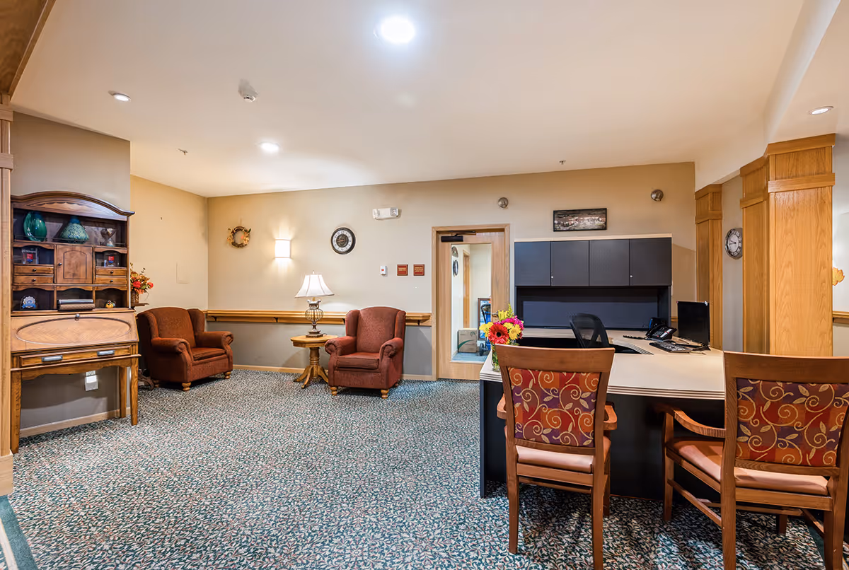 A cozy interior room with two upholstered armchairs and a small round table with a lamp between them. There is a wooden desk with two patterned chairs in the foreground, and a built-in cabinet with dark doors behind the desk. The walls are beige, and the carpet has a floral pattern. Decorative items and a clock are mounted on the walls.