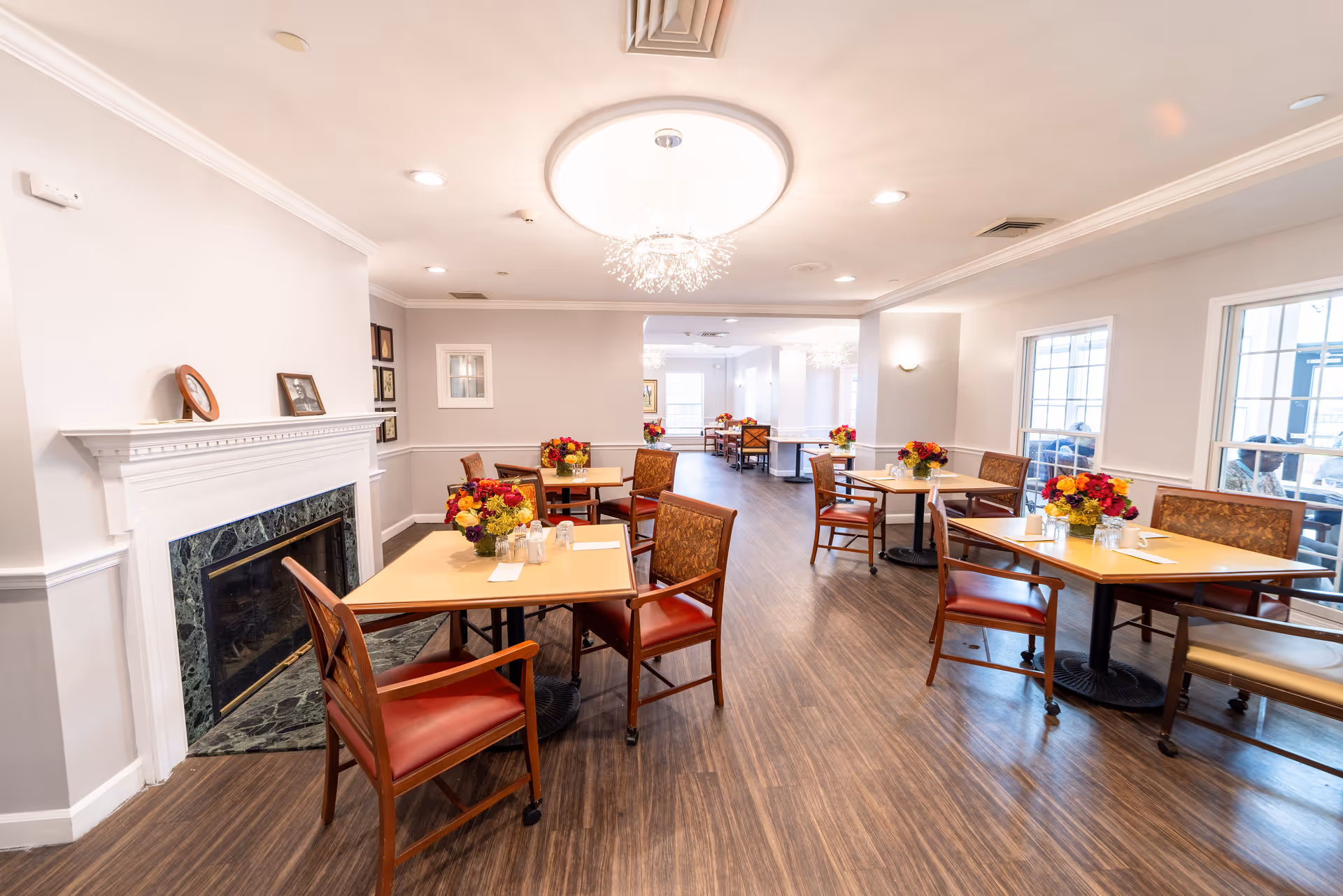 A bright dining room with multiple tables and chairs arranged neatly. Each table has a colorful flower arrangement. The room features a white fireplace with a green marble surround on the left side and large windows on the right side letting in natural light. The ceiling has modern light fixtures and recessed lighting.