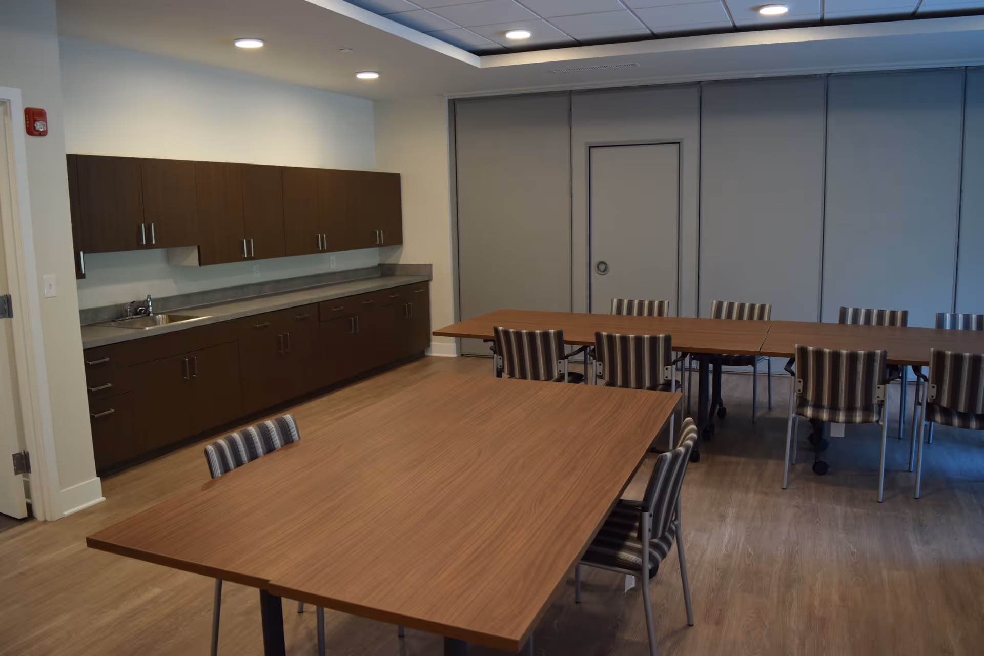 A room with multiple wooden tables and striped chairs arranged around them. Along one wall, there are dark brown cabinets with a countertop and a small sink. The room has a light wood floor and a ceiling with recessed lighting. The far wall has a closed door and large gray panels.