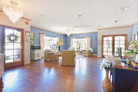 A bright and spacious living room area with wooden flooring, light blue walls, and large windows letting in natural light. The room is furnished with comfortable armchairs and sofas arranged around a coffee table. There is a wooden door with glass panels on the left and another set of glass doors on the right. Decorative elements include a chandelier, table lamps, framed pictures, and a vase with flowers on a wooden cabinet.