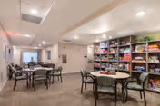 A well-lit common area with multiple round tables and chairs arranged for group activities or socializing. One wall features shelves filled with board games and puzzles. The space has a neutral color palette with beige walls and carpeted flooring.