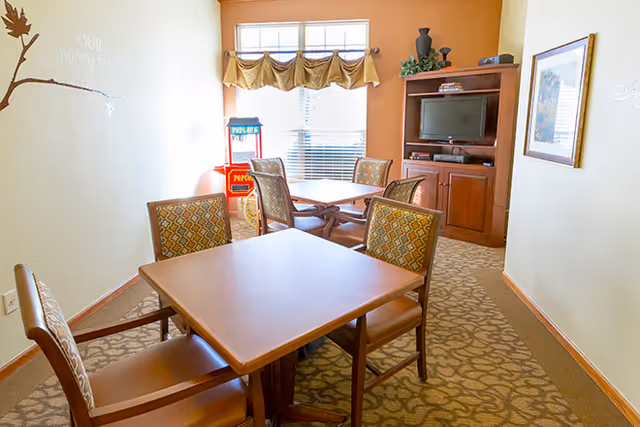 A small common area with two square tables surrounded by patterned chairs. A popcorn machine is placed near a window with beige curtains. A wooden entertainment center with a TV and decorative items is against the far wall. The room has beige walls and carpet with a floral pattern.