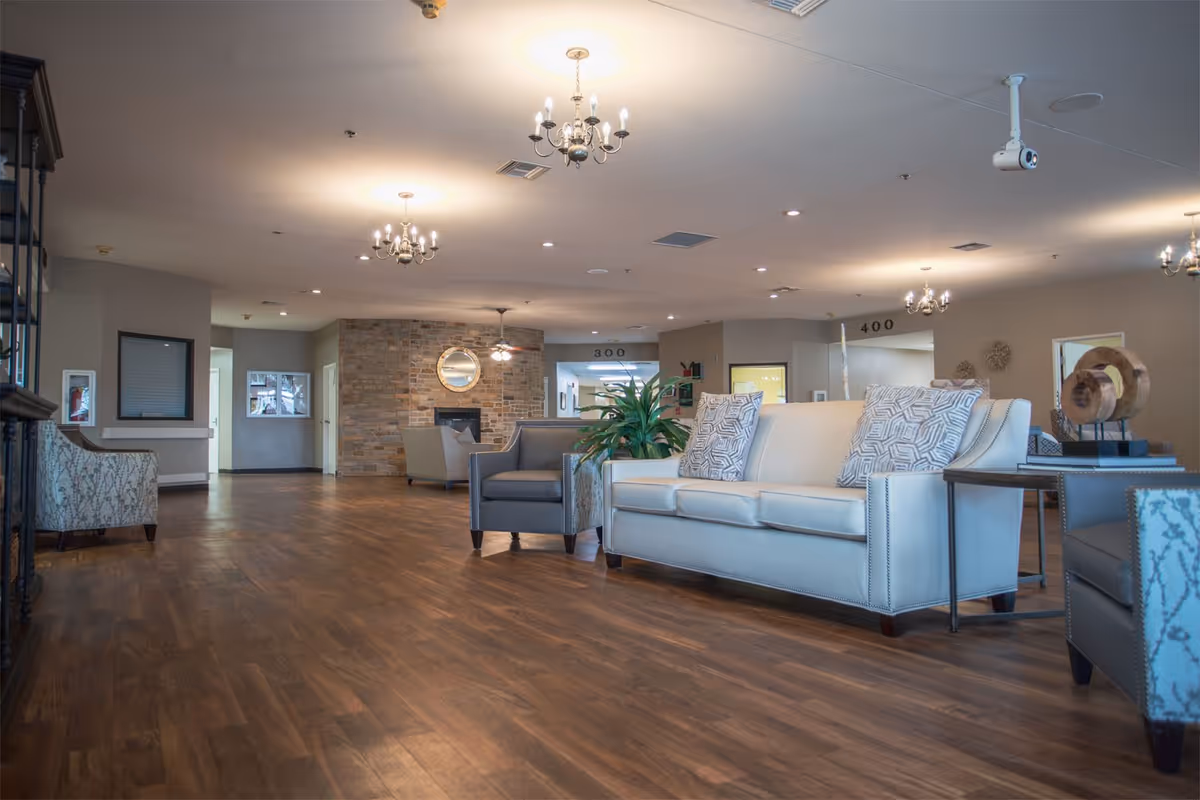 Spacious nursing home lobby with sofas, armchairs, chandeliers, wooden floor and a stone fireplace in the background.