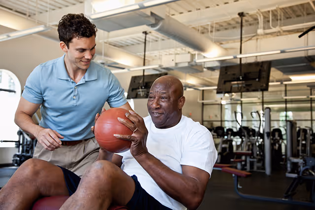 An older man in a white t-shirt holding a medicine ball while sitting on a bench in a gym, assisted by a younger man in a light blue polo shirt who is smiling and supporting him.