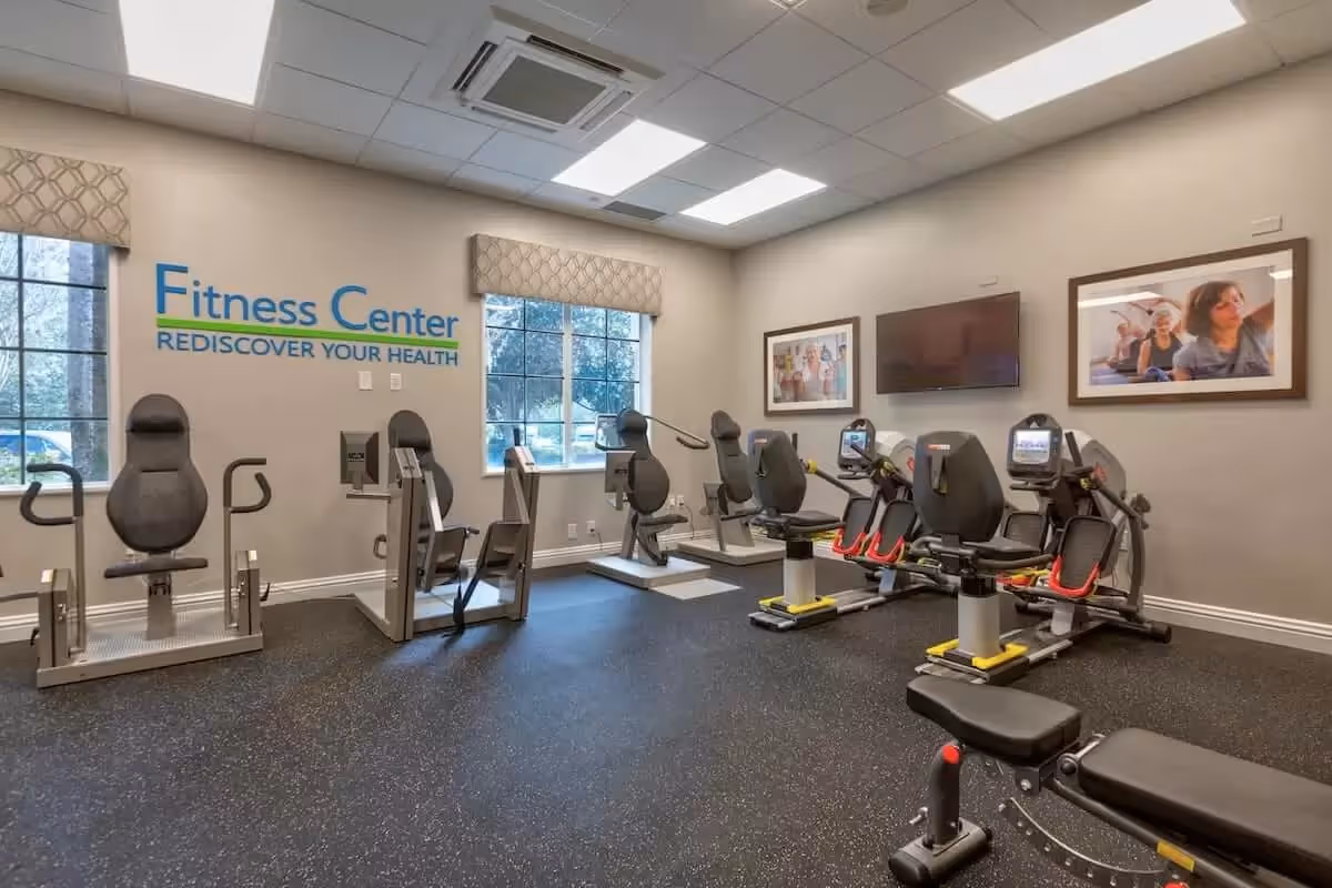 A fitness center room with multiple recumbent exercise machines, a bench, windows, framed photos and a wall-mounted TV.