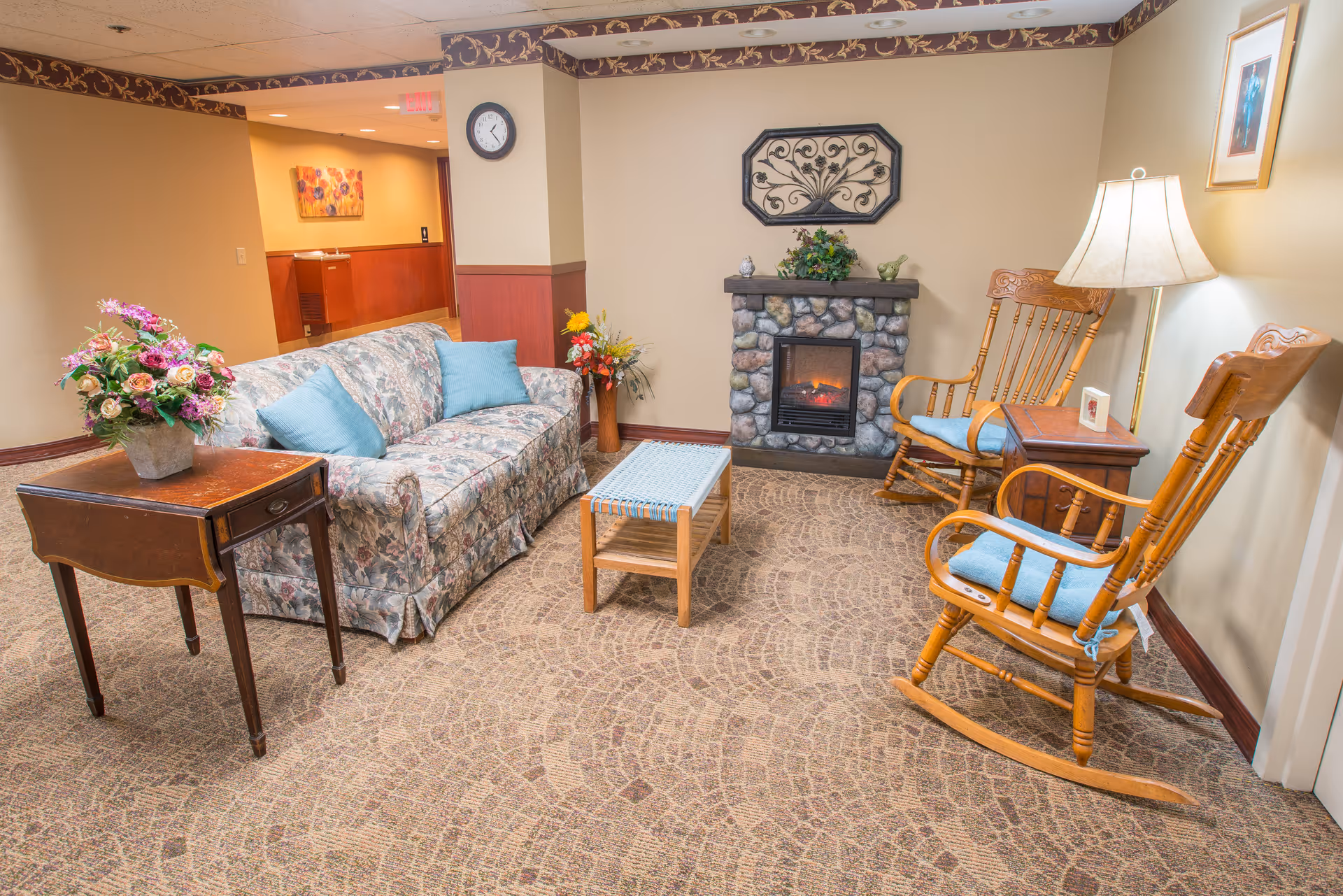 A cozy sitting area with a floral sofa, two wooden rocking chairs, a small electric fireplace and floral arrangements.