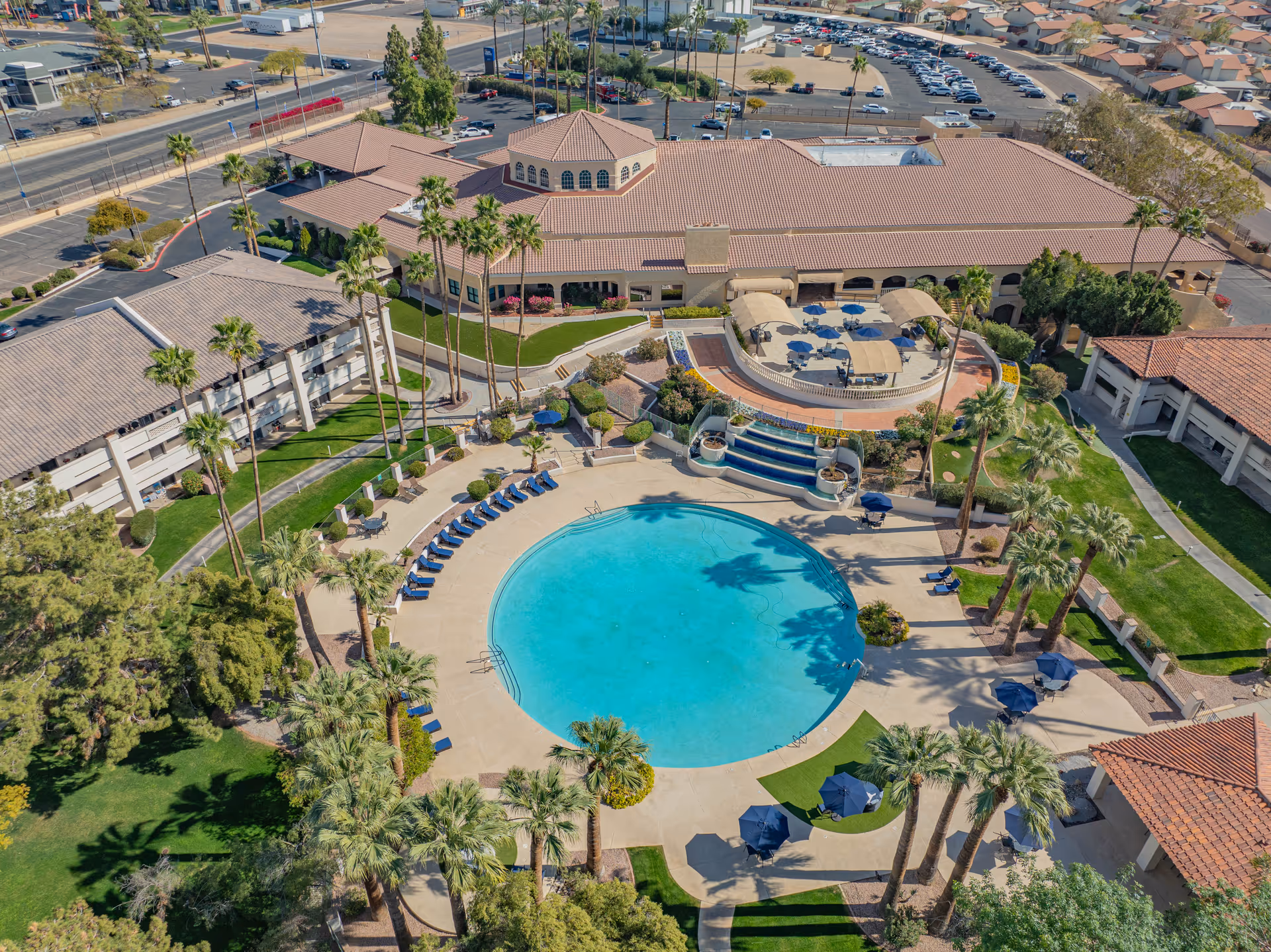 Aerial view of a senior living campus with a large circular swimming pool surrounded by lounge chairs, palm trees, and Mediterranean-style buildings.