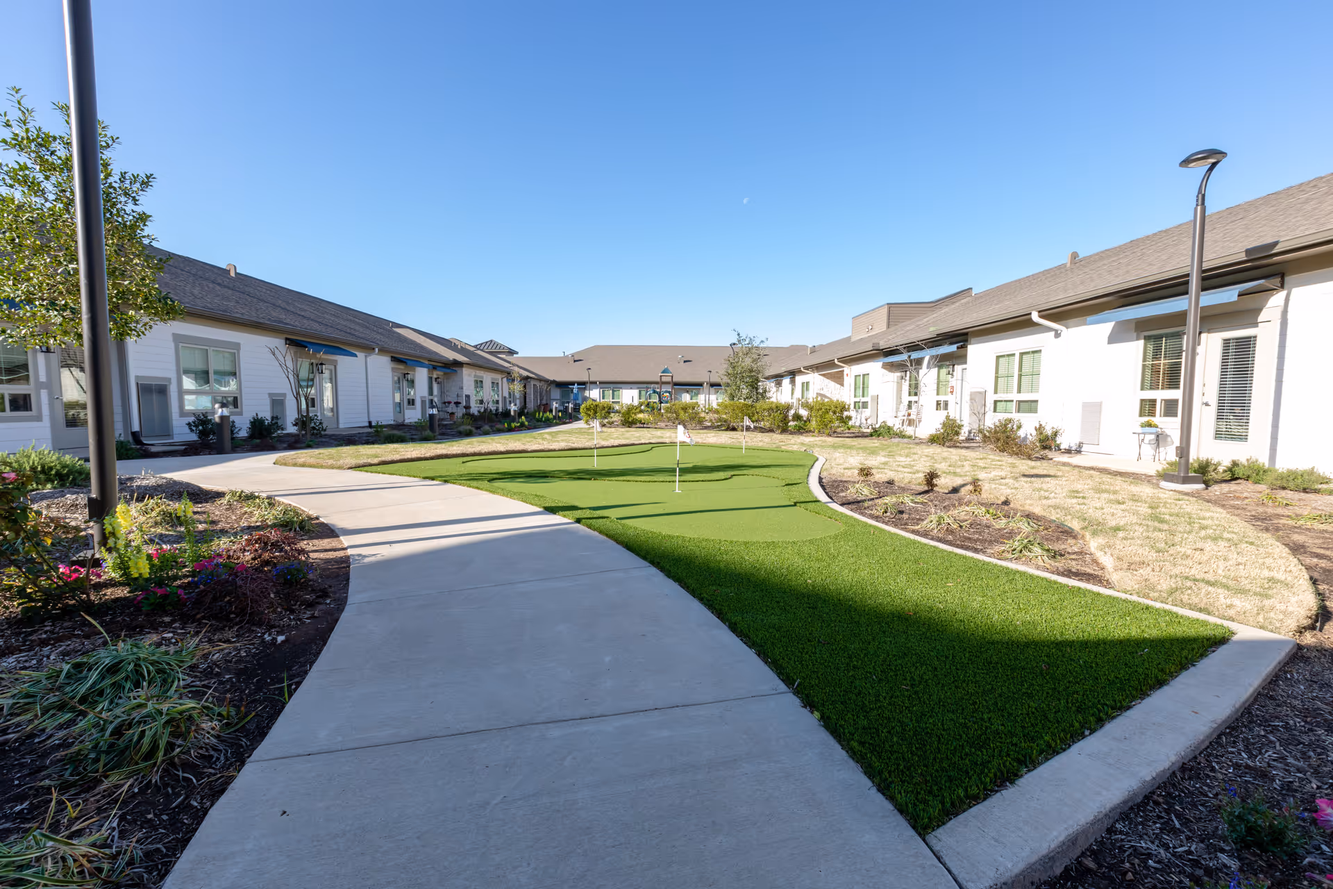 Outdoor courtyard area at The Village at Sugar Land featuring a putting green with three small flags, surrounded by a curved concrete walkway, landscaped flower beds, and single-story buildings with windows and awnings under a clear blue sky.