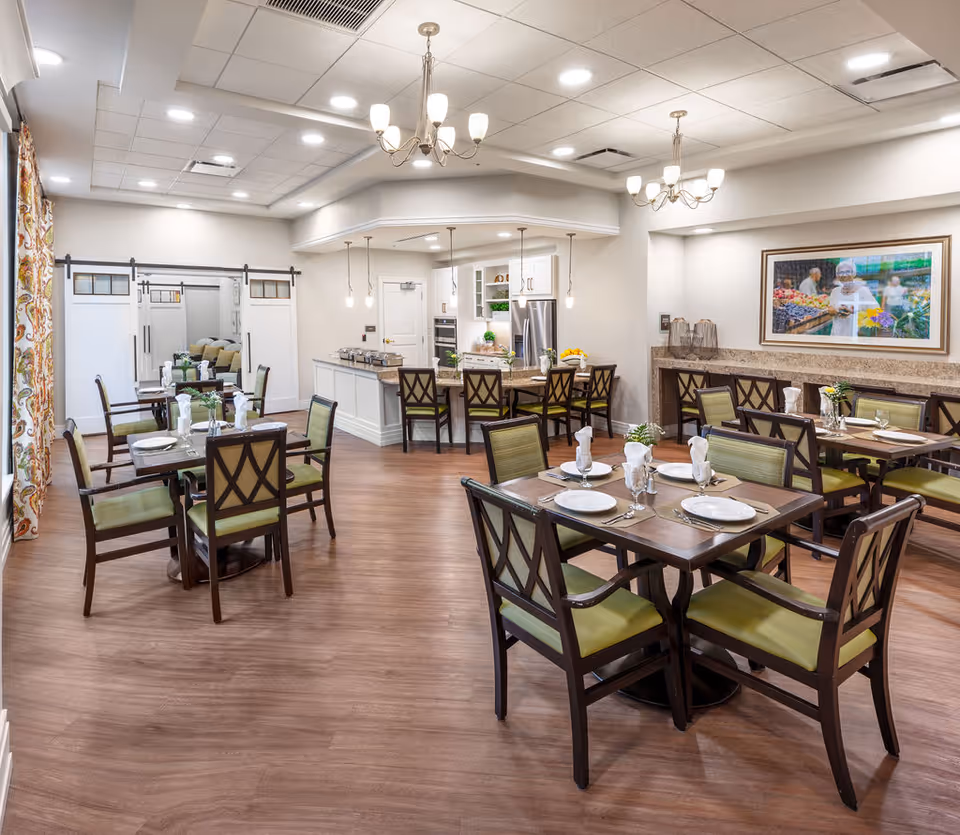 A bright and spacious dining room in a senior living facility with several wooden tables and chairs arranged neatly. Each table is set with white plates, glasses, and folded napkins. The room features wood flooring, chandeliers, and pendant lights hanging from a white ceiling. In the background, there is a kitchen area with a refrigerator, oven, and countertop. A large framed picture hangs on the wall, and sliding barn doors are visible on the left side of the room.