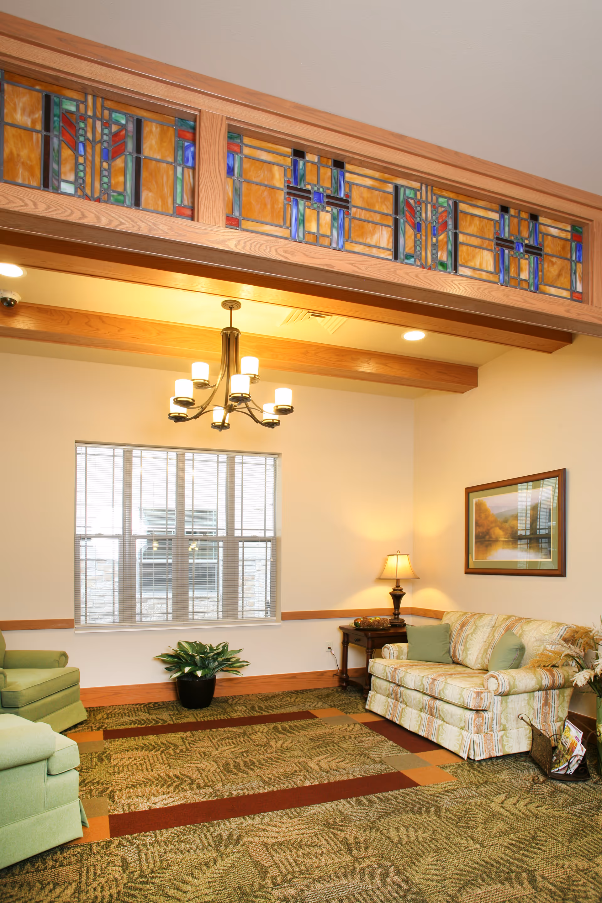 A cozy living room area with a patterned green carpet, a floral upholstered sofa with green cushions, a green armchair, a wooden side table with a lamp, a framed landscape painting on the wall, a potted plant near the window, and a decorative stained glass panel above the doorway.