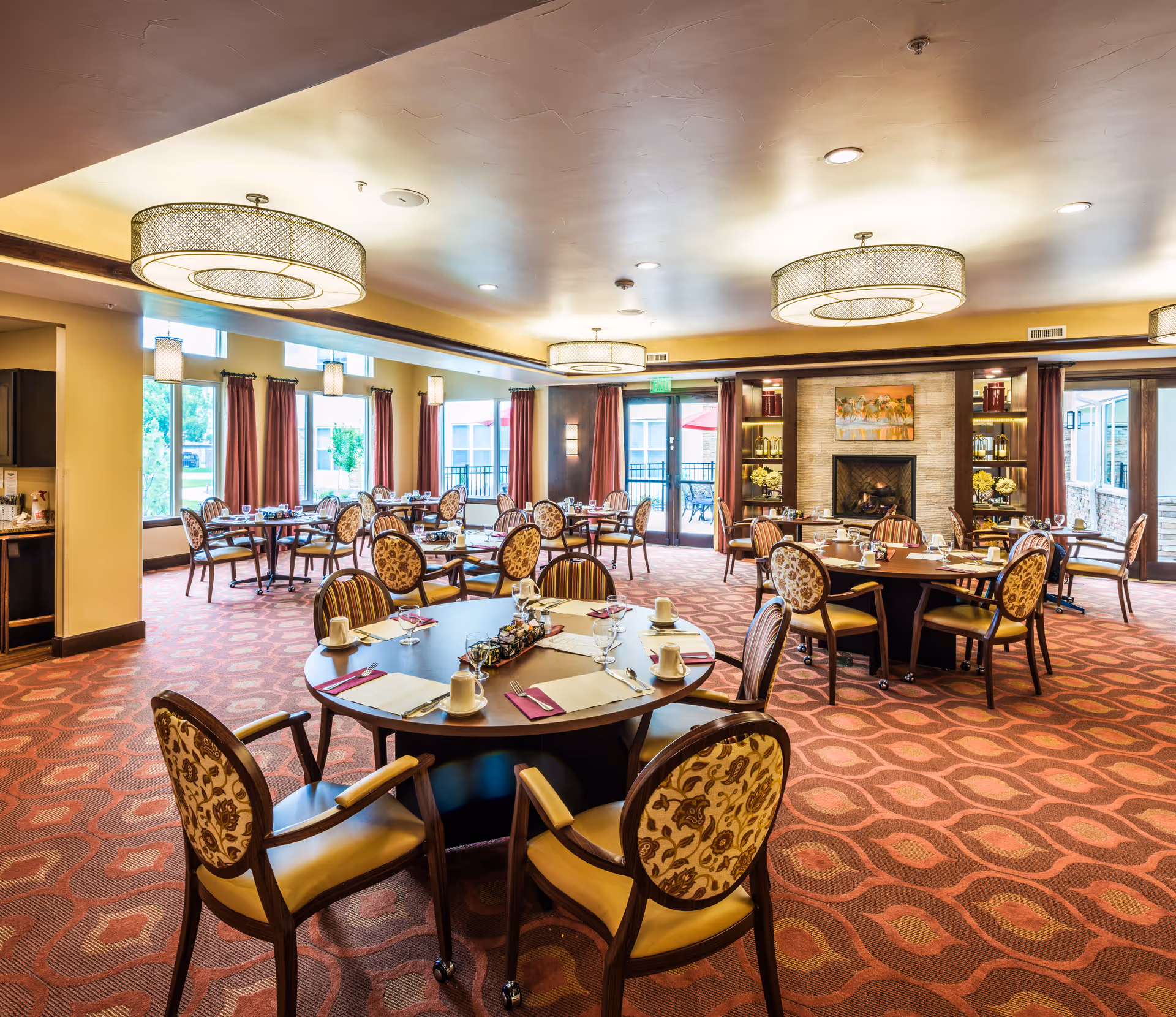 A spacious dining room in an assisted living facility with round tables set for meals. Each table has chairs with patterned upholstery, place settings with cups, glasses, and napkins. The room features large windows with red curtains, modern ceiling lights, a fireplace with decorative shelves, and doors leading to an outdoor patio area.