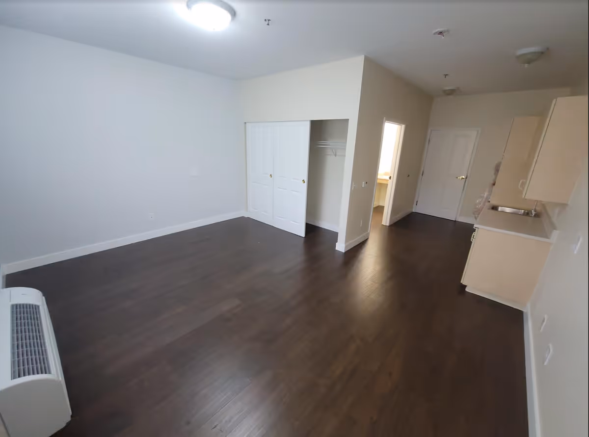 Empty studio-style interior with dark wood floors, a small kitchenette on the right, a closet with sliding doors, and doors to a bathroom and entryway.