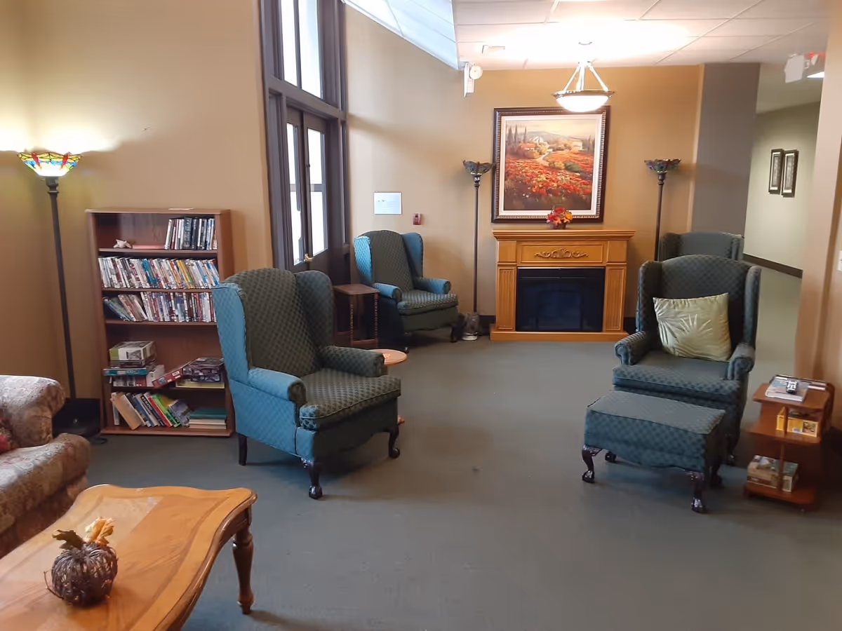 A cozy living room area in the River View facility featuring four green upholstered armchairs, a wooden coffee table with a decorative pumpkin, a wooden bookshelf filled with books and games, a wooden electric fireplace with a framed landscape painting above it, and two floor lamps with stained glass shades.
